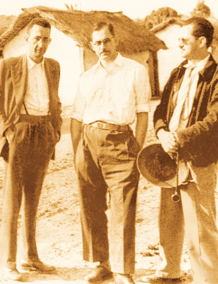 Foto mostra os três homens de pé diante de casas de barro antigas. Um deles segura um chapéu e um estetoscópio.