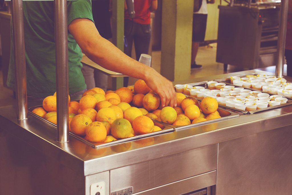 Foto com recorte mostra uma pessoa segurando uma bandeja e escolhendo um laranja em um bancada com diversas laranjas e pequenos copos plásticos com um creme na cor laranja. Ao fundo, há pessoas em pé de frente para outras bancadas com comida. 