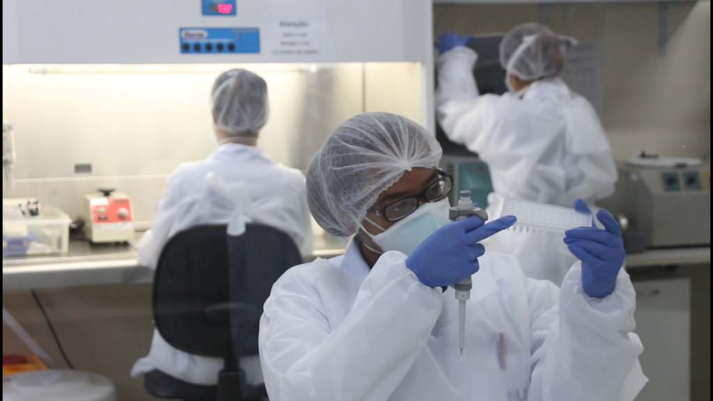 Foto de uma funcionária devidamente equipamda analisando uma amostra. Laboratório de Genética e Biologia Molecular (LGBM) do Nupad. Imagem: TV UFMG / UFMG